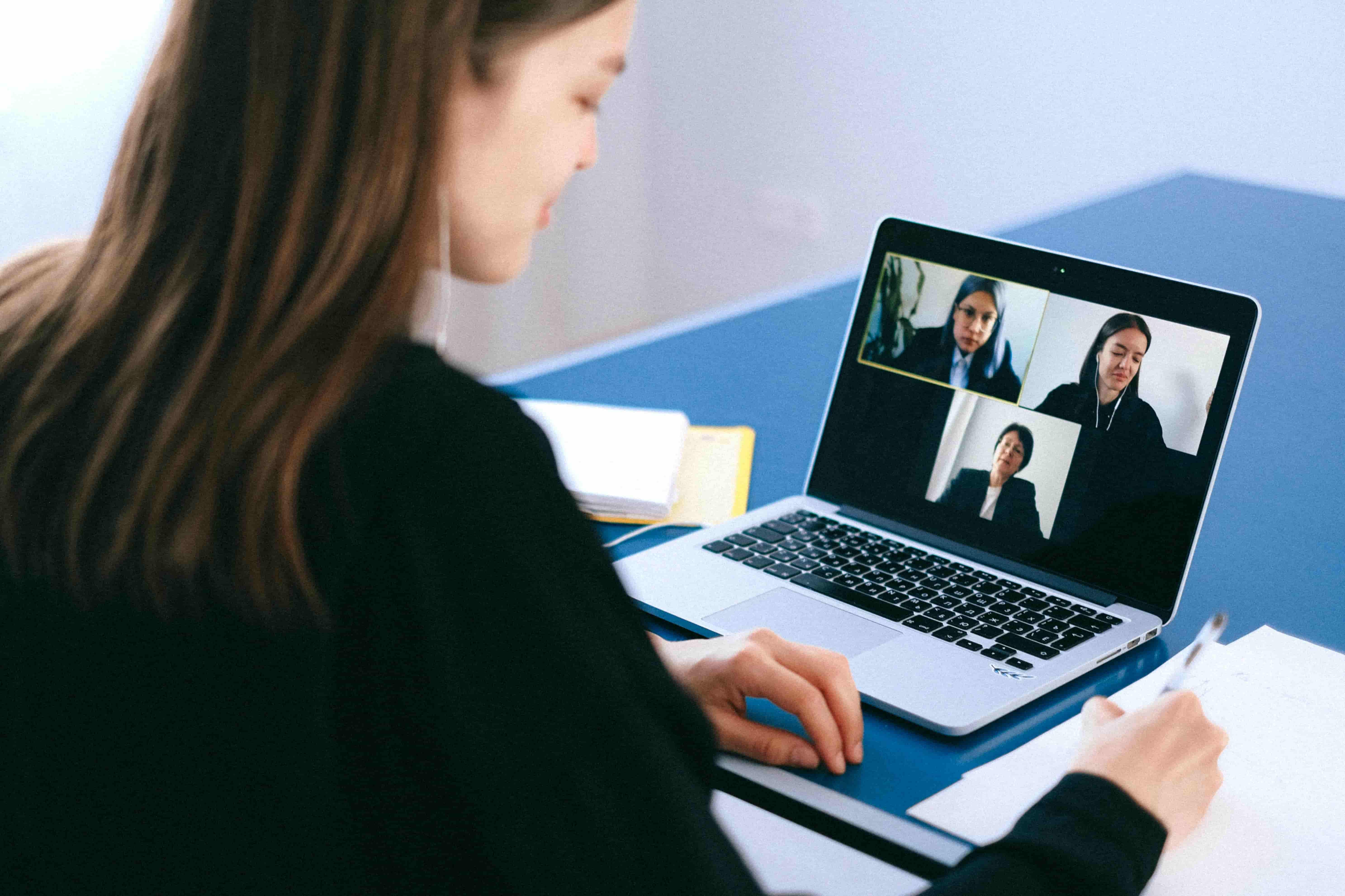 Woman on a laptop video zoom call with three people, taking notes on paper at a desk.
