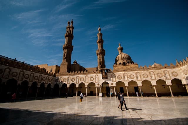 A wide courtyard of an historic mosque under a clear blue sky, featuring two prominent minarets and a large central dome. The marble courtyard is surrounded by arched colonnades with intricate geometric patterns, and a few people are walking or standing in the open space.