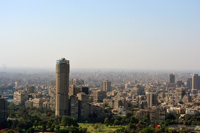 A wide view of Cairo, Egypt, showcasing a dense urban skyline with high-rise buildings and a prominent tall cylindrical tower surrounded by greenery in the foreground.