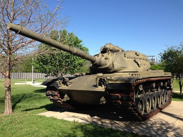 A military tank displayed outdoors on a concrete platform surrounded by grass and trees, under a clear blue sky.