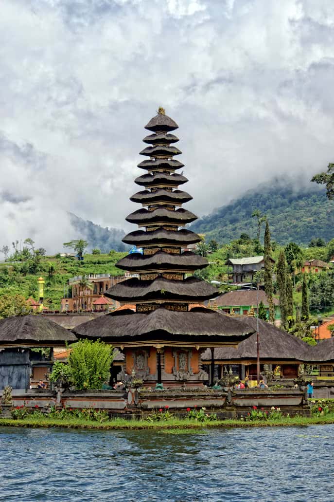 A multi-tiered pagoda with traditional straw roofing sits by a lake, surrounded by lush greenery and misty mountains in the background.