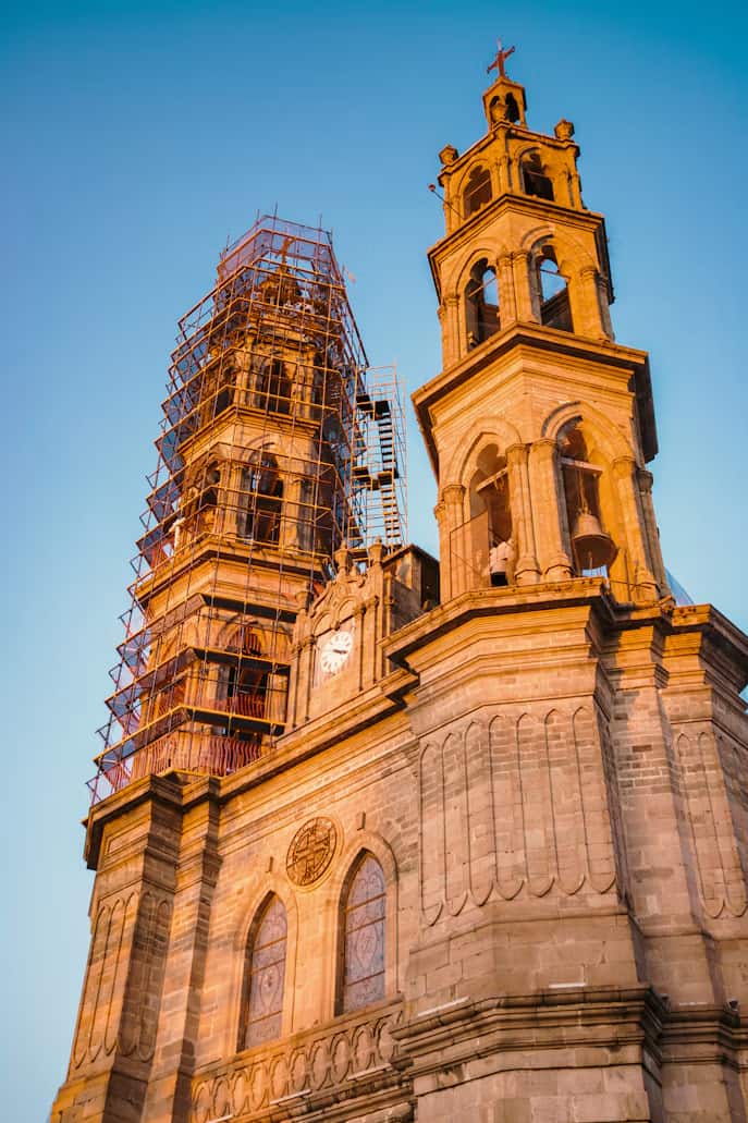 Stone cathedral with two tall towers, one under renovation with scaffolding, in warm sunlight against a clear blue sky.