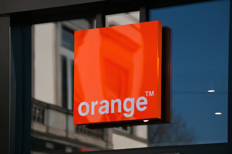 Bright orange sign with "orangeTM™" in white, displayed on a storefront window reflecting nearby buildings.