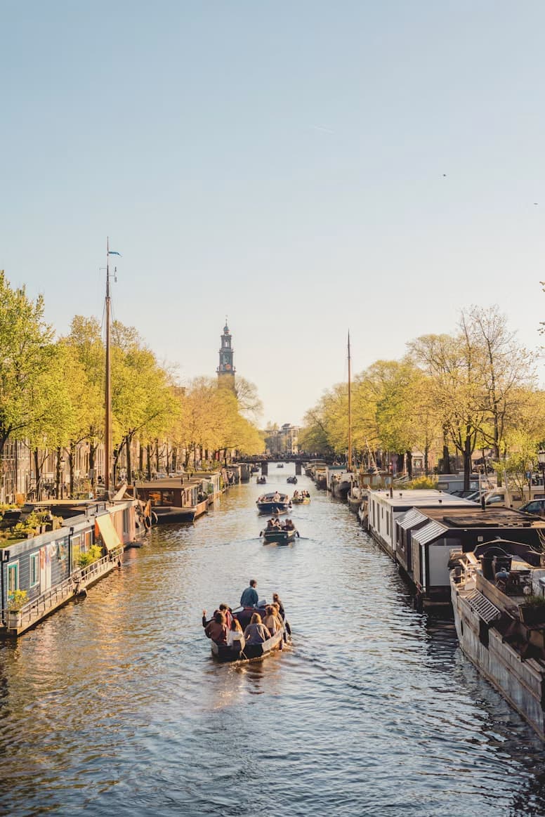 People in boats on a canal in a city with houseboats and trees lining the waterway, under a clear blue sky.