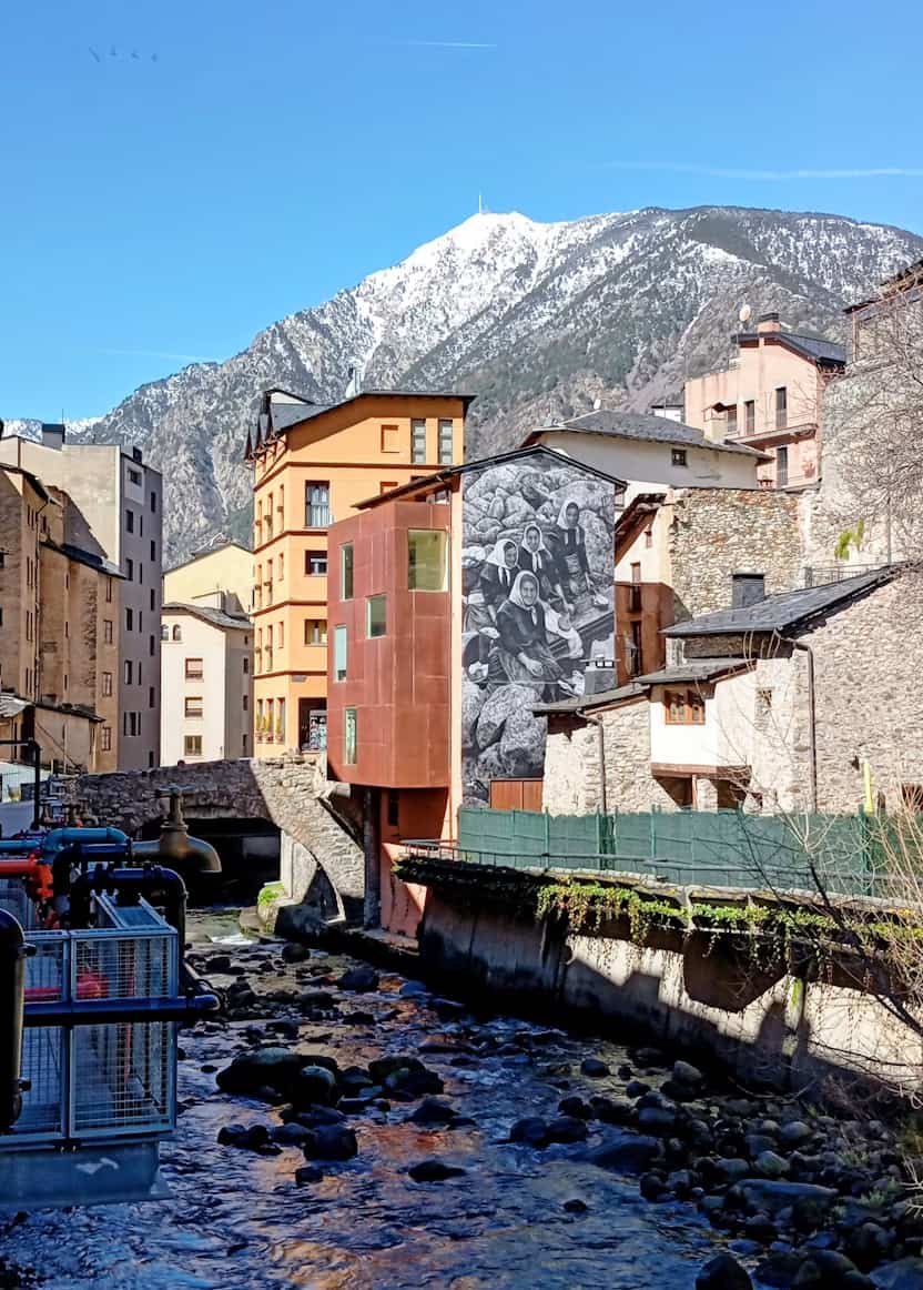 A picturesque mountain town with colorful buildings, a mural on one wall, a rocky stream, and snow-capped mountains in the background under a clear blue sky.