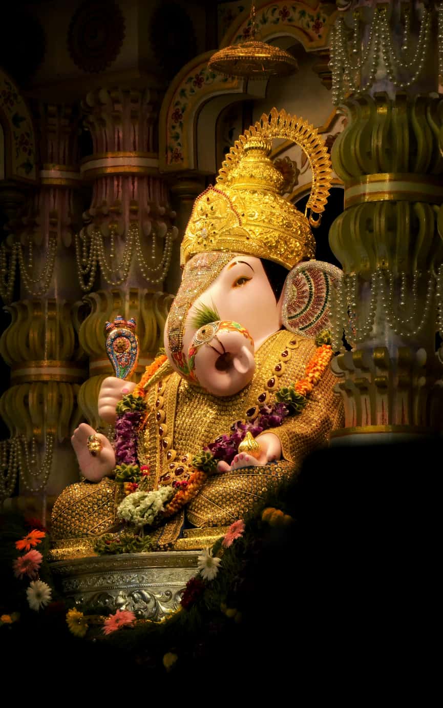 A richly adorned statue of Lord Ganesha, wearing a golden crown and garlands, with intricate details and vibrant colors, against a decorative backdrop.