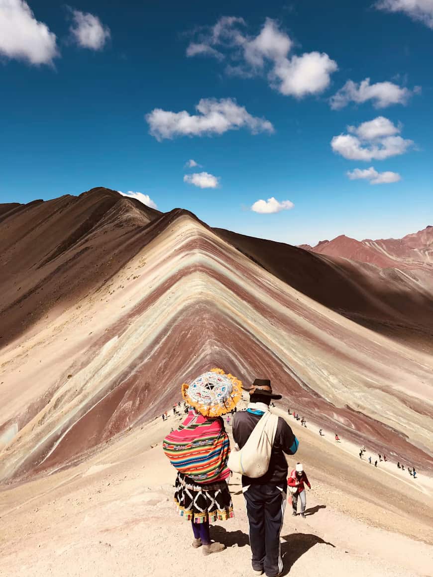 Two people in colorful attire stand on a trail overlooking the multicolored Rainbow Mountain under a bright blue sky with scattered clouds.