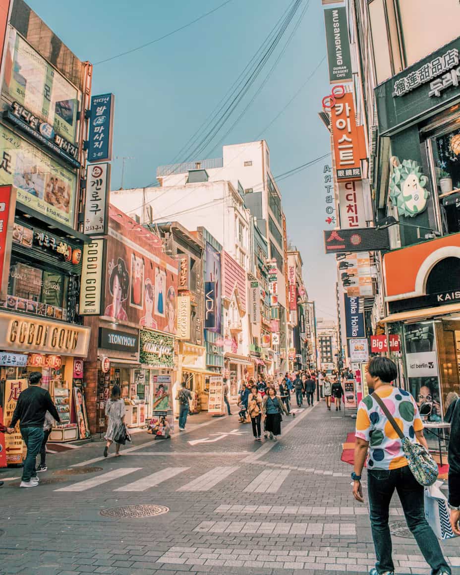 Bustling street in a city with bright signs and shops, people walking on the sidewalk beneath a clear blue sky.