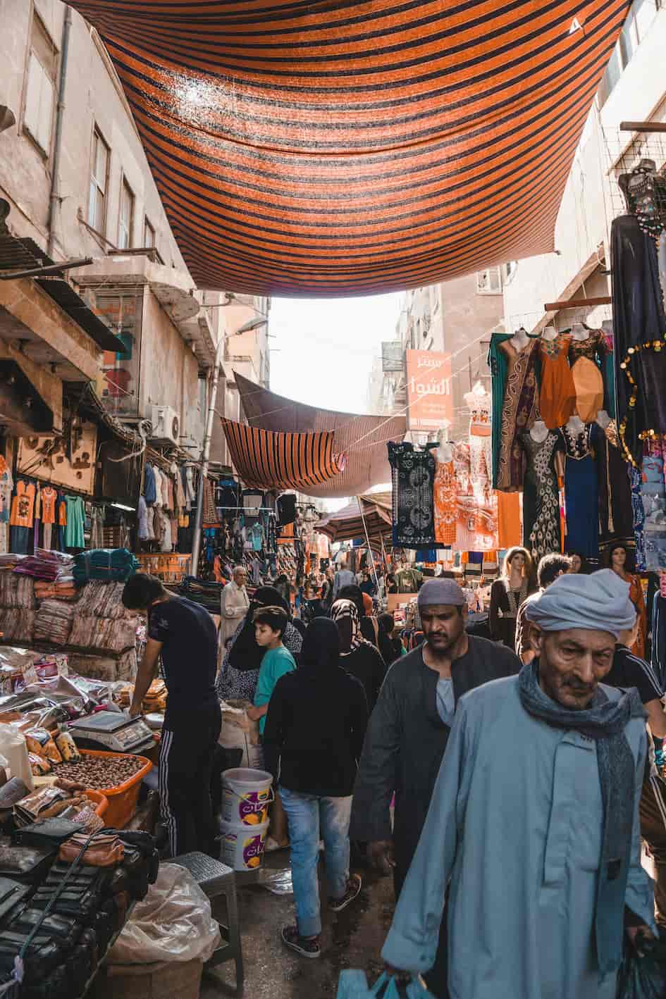 A bustling outdoor market with vendors selling clothes and goods, shaded by striped awnings, and crowded with people walking and shopping.