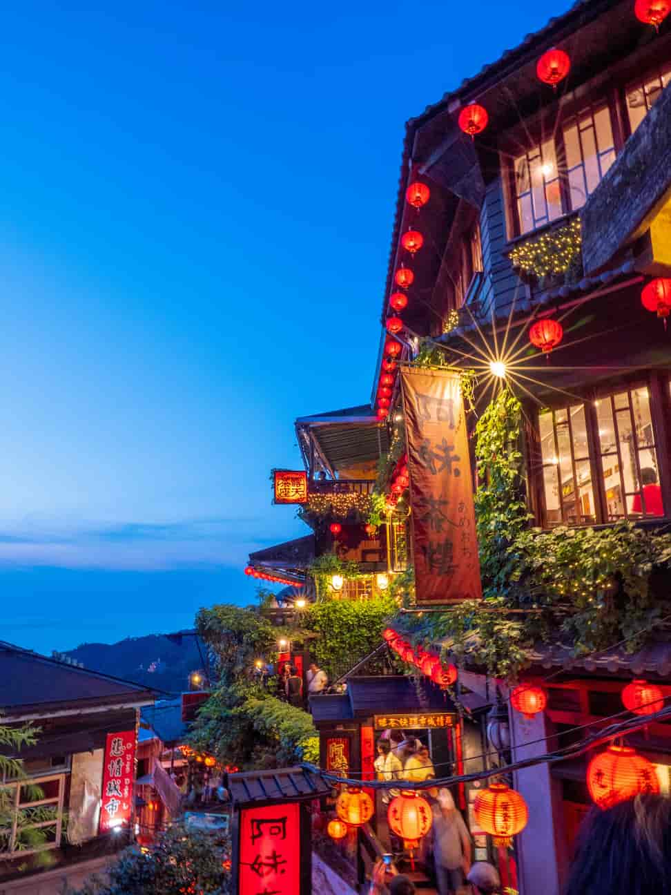 A vibrant street scene at dusk with illuminated red lanterns, colorful signs, and ivy-covered buildings against a blue sky.